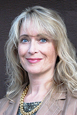 White woman with blonde wave hair with bangs. Wearing a brown jacket and gold necklace